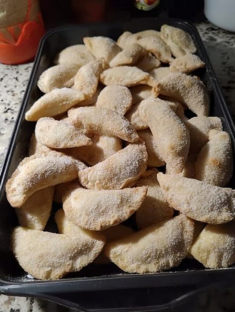 Empanaditas de queso crema y dulce de leche espolvoreadas con azúcar en charola negra sobre encimera de cocina.