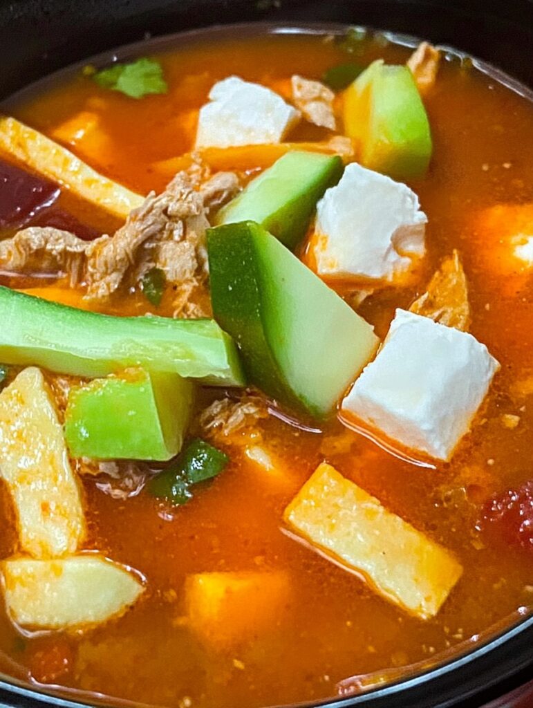 Tazón de caldo tlalpeño con aguacate, queso fresco, tiras de tortilla frita y pollo deshebrado en caldo rojo.