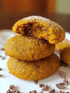 Tres galletas de calabaza y chía sin harina ni azúcar, apiladas y espolvoreadas ligeramente, con trozos de chocolate alrededor.